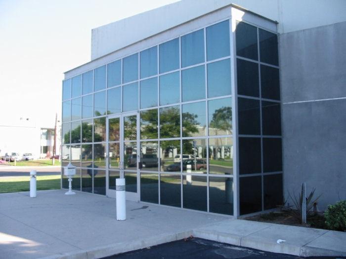 Budynek biurowy z częściowo zaciemnionymi szybami, widoczne odbicie drzew i samochodów.