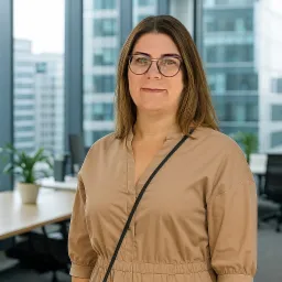 Kobieta w okularach, ubrana w beżowy kombinezon, stoi w nowoczesnym biurze na tle przeszklonych ścian i biurek. Delikatny uśmiech, profesjonalny wygląd.