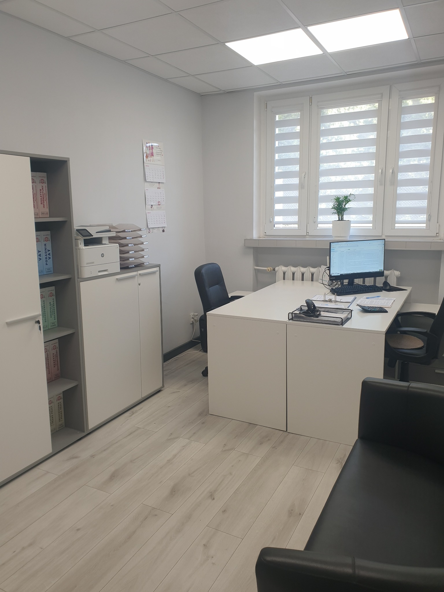 Jasne biuro z biurkiem, komputerem, regałem z segregatorami i kalendarzem na ścianie. Widok na okno z żaluzjami. Wnętrze biura rachunkowego.