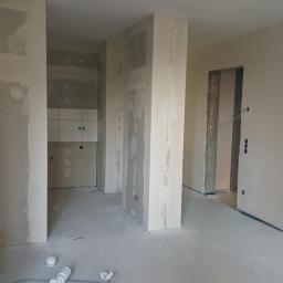 Krystian Głuc - Wnętrze w trakcie prac wykończeniowych, widoczne ślady gładzi na ścianach i kolumnach, przygotowane punkty elektryczne i rury na podłodze, w tle fragment kuchni z białymi płytkami.