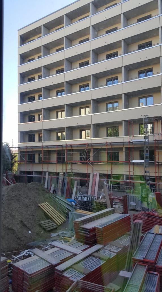 Wielopiętrowy budynek mieszkalny w trakcie budowy lub remontu, z widocznymi rusztowaniami, stertami materiałów budowlanych i otwartymi balkonami.