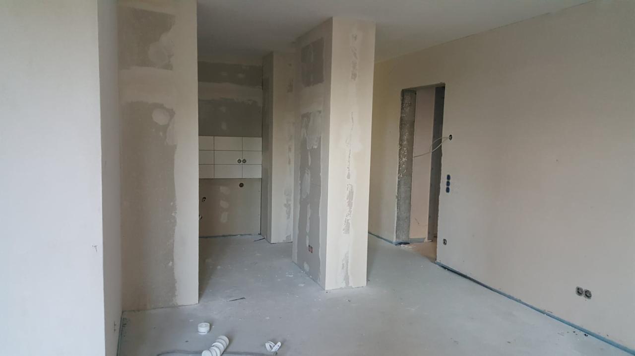 Wnętrze w trakcie prac wykończeniowych, widoczne ślady gładzi na ścianach i kolumnach, przygotowane punkty elektryczne i rury na podłodze, w tle fragment kuchni z białymi płytkami.