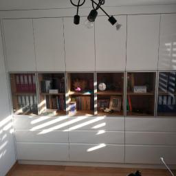 Awan Meble - Biała zabudowa ścienna z szafkami górnymi, częściowo przeszklonymi, i dolnymi szufladami, widoczne segregatory, książki i dekoracje, oświetlenie sufitowe z żarówkami.