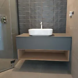 Szara szafka łazienkowa z blatem i umywalką nablatową w nowoczesnej łazience z szarymi płytkami o fakturze 3D, widoczna kabina prysznicowa ze szklanymi drzwiami.