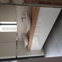 Awan Meble - Łazienka w trakcie wykańczania z dwiema umywalkami nablatowymi na drewnianym blacie i białymi szafkami pod nimi, widoczne fragmenty surowego muru.