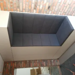 Wnęka z siedziskiem tapicerowanym szarą tkaniną, obudowana białymi płytami meblowymi, na tle ceglanej ściany i szarej podłogi w trakcie remontu.