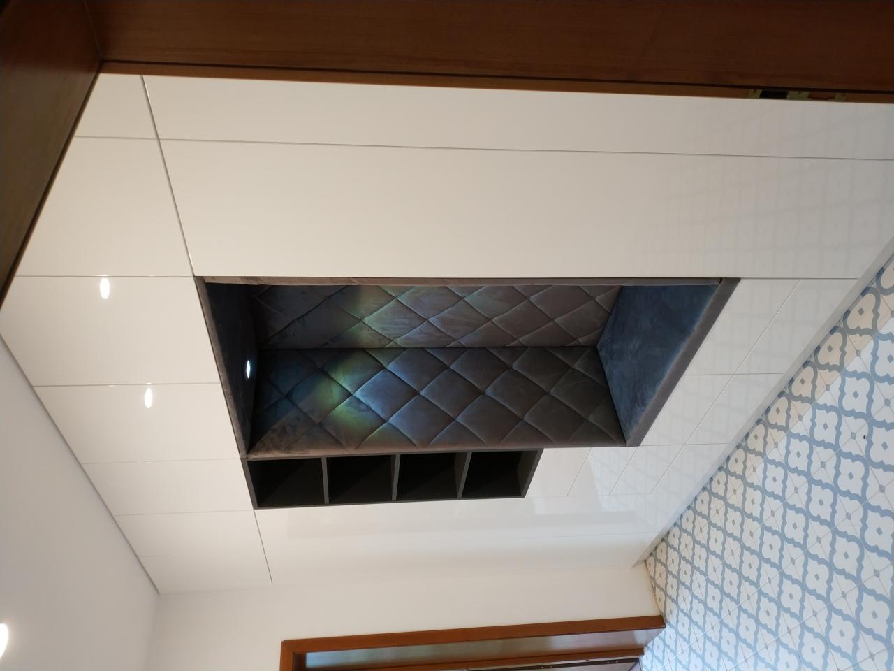 Biała zabudowa meblowa z wnęką wykończoną tapicerowanym panelem w romby i półkami, na tle wzorzystej podłogi.