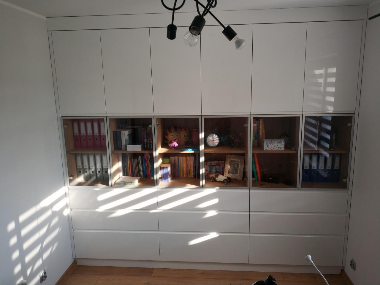 Biała zabudowa ścienna z szafkami górnymi, częściowo przeszklonymi, i dolnymi szufladami, widoczne segregatory, książki i dekoracje, oświetlenie sufitowe z żarówkami.