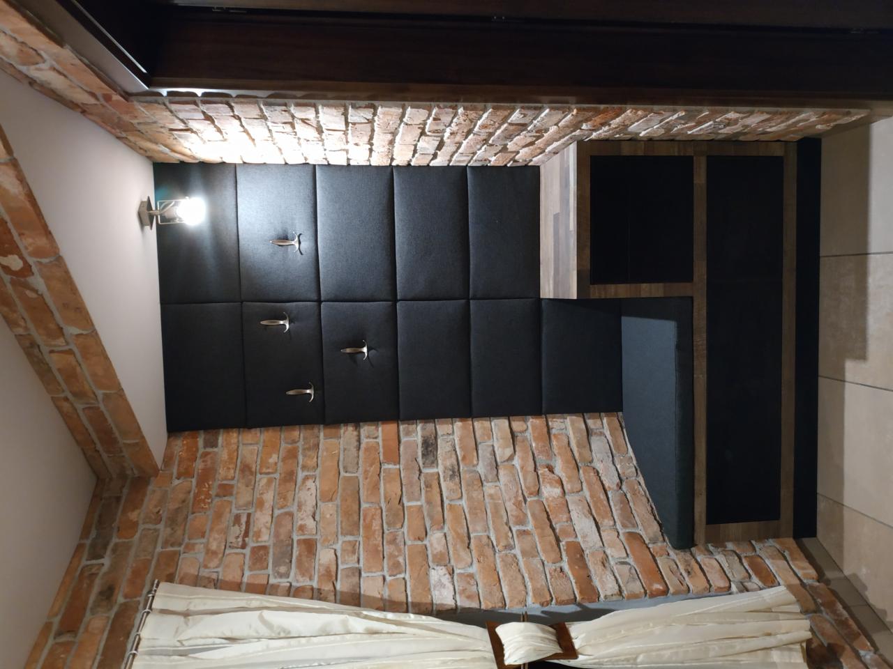 Czarny tapicerowany zagłówek łóżka z metalowymi wieszakami, ceglana ściana, oświetlenie ścienne i zasłony w sypialni.