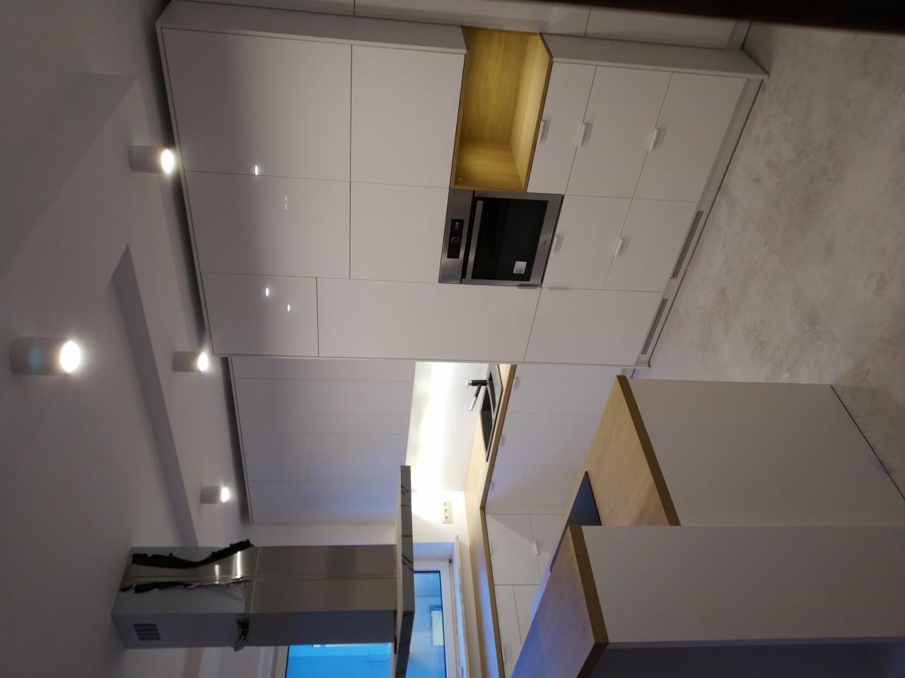 Nowoczesna kuchnia z białymi, lakierowanymi frontami szafek, drewnianymi akcentami wewnątrz otwartych półek i na wyspie kuchennej, oświetleniem punktowym w suficie i minimalistycznym okapem.