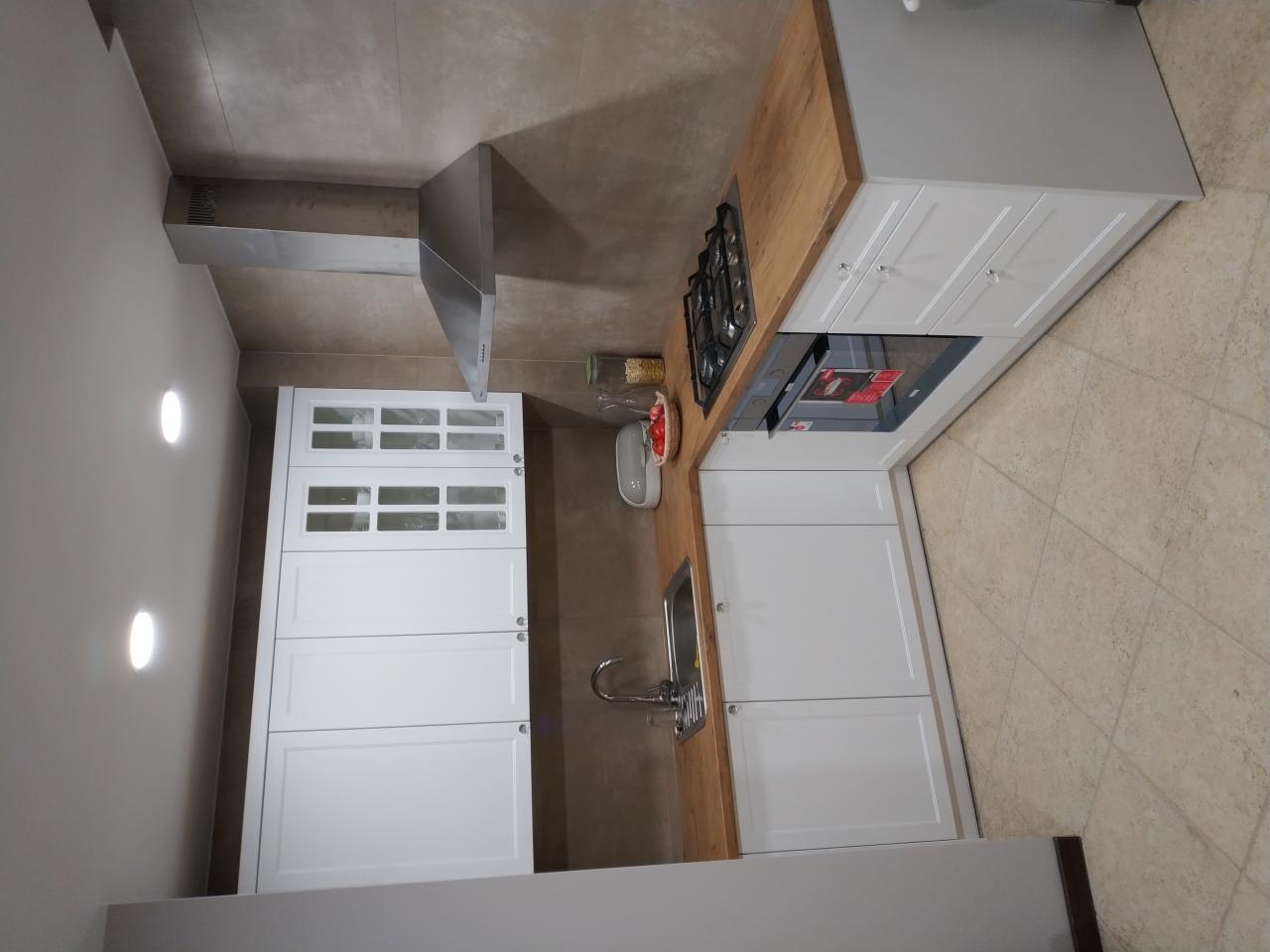 Nowoczesna kuchnia z białymi szafkami, drewnianym blatem i srebrnym okapem, widok na płytę gazową i szklane fronty szafek wiszących.