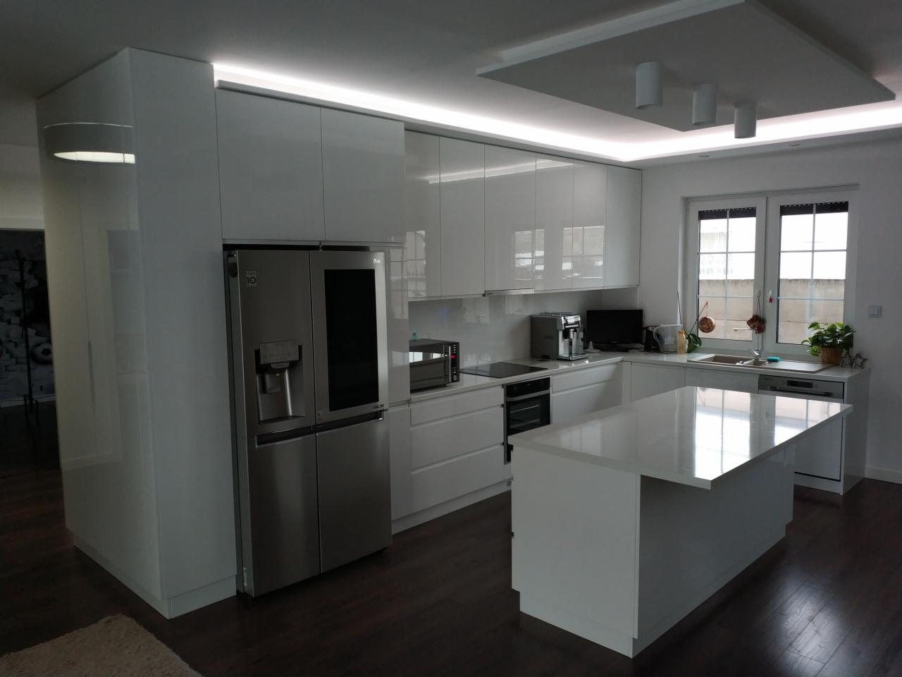 Nowoczesna kuchnia w bieli z wyspą, zabudową po sufit i oświetleniem LED podszafkowym oraz dużym, srebrnym lodówko-zamrażarką.