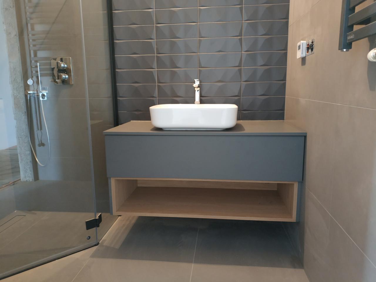 Szara szafka łazienkowa z blatem i umywalką nablatową w nowoczesnej łazience z szarymi płytkami o fakturze 3D, widoczna kabina prysznicowa ze szklanymi drzwiami.