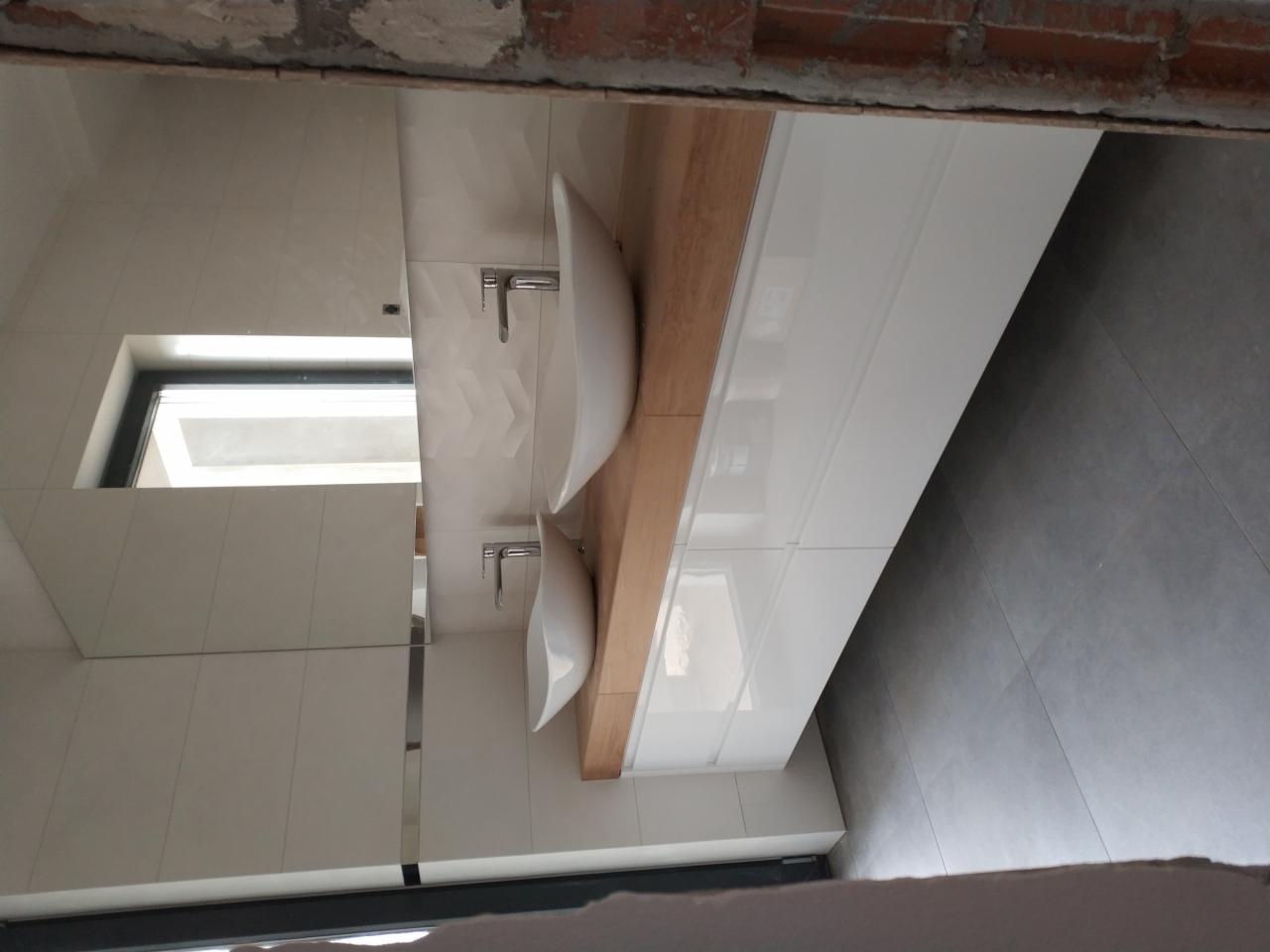 Łazienka w trakcie wykańczania z dwiema umywalkami nablatowymi na drewnianym blacie i białymi szafkami pod nimi, widoczne fragmenty surowego muru.