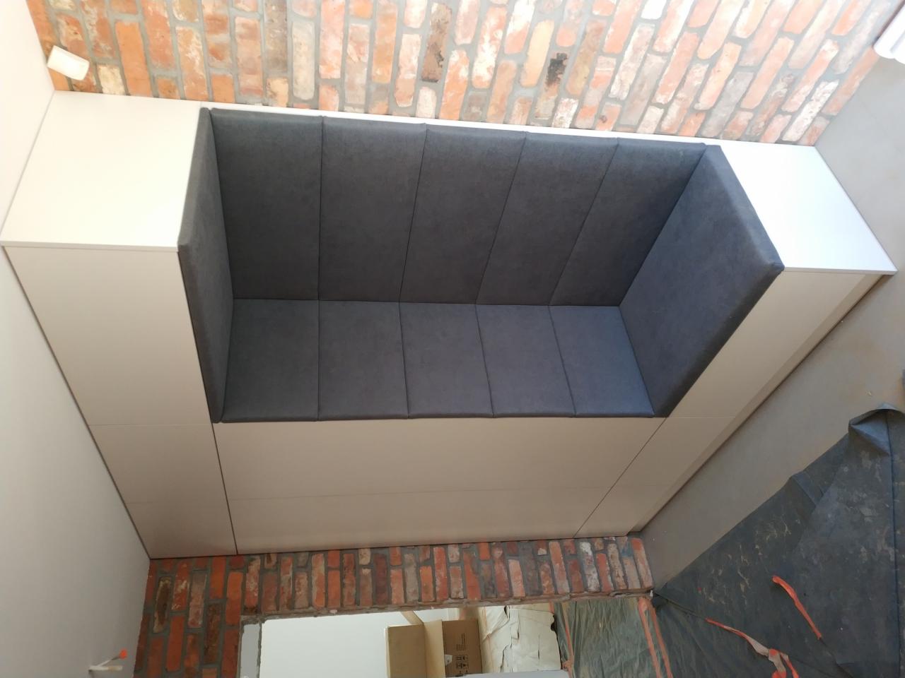 Wnęka z siedziskiem tapicerowanym szarą tkaniną, obudowana białymi płytami meblowymi, na tle ceglanej ściany i szarej podłogi w trakcie remontu.