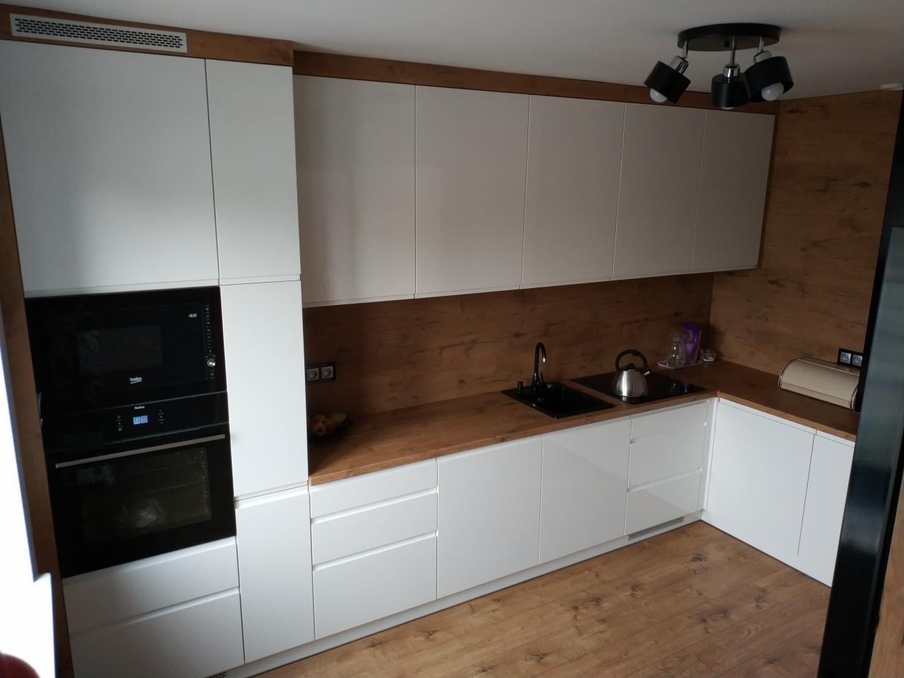 Nowoczesna kuchnia z białymi, lakierowanymi frontami szafek, drewnianym blatem i panelem ściennym, czarnym zlewem i czajnikiem na blacie, oświetlona reflektorami sufitowymi.