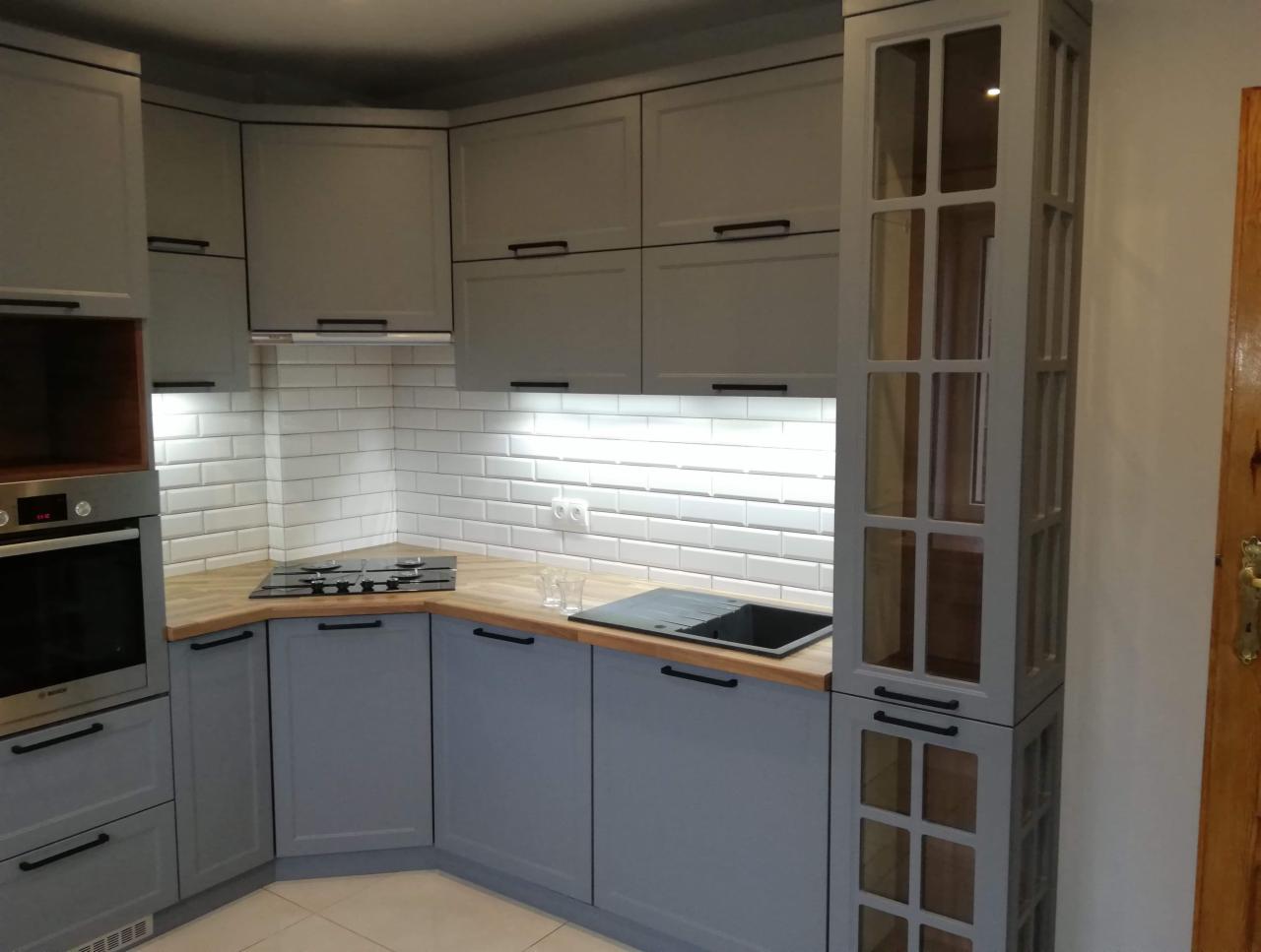 Szare szafki kuchenne z drewnianym blatem i białymi płytkami metra, widoczne płyty grzewcze i zabudowana kolumna z przeszklonymi drzwiczkami.