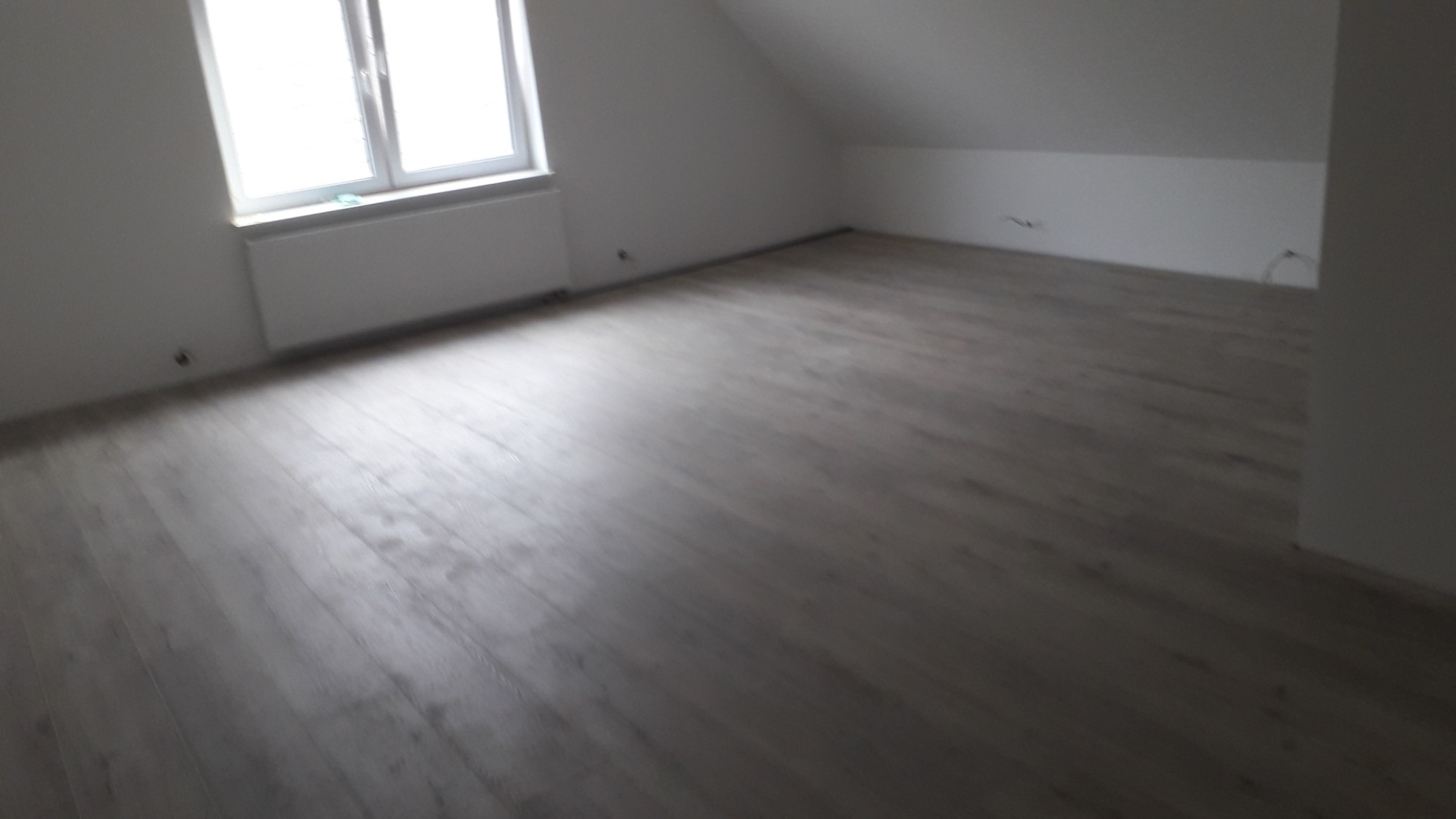 Jasnoszara podłoga z paneli w pokoju na poddaszu, widok na okno i grzejnik.