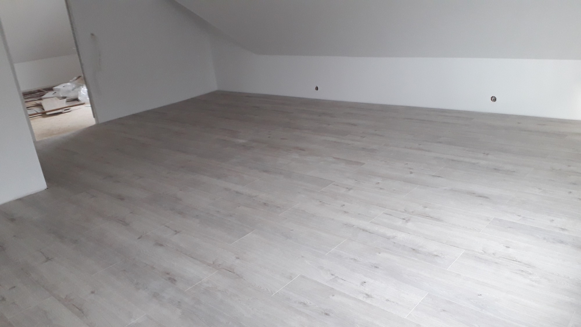 Jasne panele podłogowe ułożone w pomieszczeniu ze skośnym sufitem, widoczne przygotowania w sąsiednim pokoju.