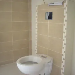 Beżowe płytki ceramiczne ułożone w łazience, widoczna toaleta i fragment kabiny prysznicowej, z niezamaskowanym otworem na gniazdko elektryczne.