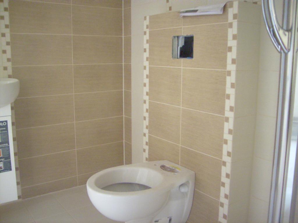 Beżowe płytki ceramiczne ułożone w łazience, widoczna toaleta i fragment kabiny prysznicowej, z niezamaskowanym otworem na gniazdko elektryczne.