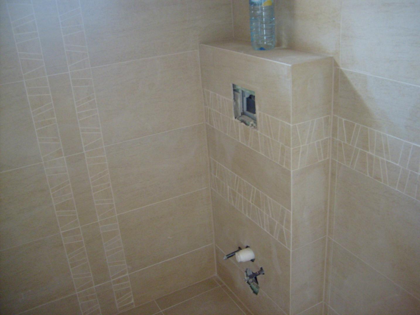 Beżowe płytki ścienne z dekoracyjnym paskiem, widoczny otwór na gniazdko i uchwyt na papier toaletowy z resztkami kleju, butelka wody na górze zabudowy.