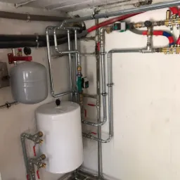 Instalacja grzewcza w piwnicy z dwoma zbiornikami, rurami miedzianymi i zaworami, widoczne elementy zabezpieczające i sterujące.