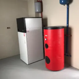 Nowoczesna instalacja pompy ciepła w kotłowni z szarymi płytkami, obejmująca biało-czarną pompę ciepła ONE, czerwony zbiornik buforowy z czarnymi zaślepkami i niebieski zbiornik wyrównawczy...