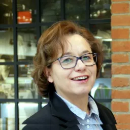 Uśmiechnięta kobieta w okularach i marynarce, z ceglaną ścianą i oknem w tle.