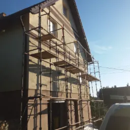 Dom jednorodzinny w trakcie prac elewacyjnych, z widocznymi rusztowaniami i częściowo odnowioną fasadą z poziomymi pasami w dwóch odcieniach brązu.