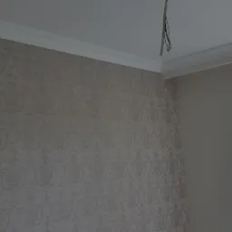 Ściana pokryta tapetą w delikatny, ornamentowy wzór, widoczna listwa dekoracyjna pod sufitem i fragment wystającego okablowania.