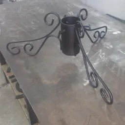 Czarny, metalowy stojak choinkowy z ozdobnymi, zakręconymi nogami, umieszczony na roboczym stole w warsztacie, widoczne ślady po obróbce metalu.