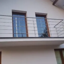 Balkon z balustradą ze stali nierdzewnej, widok z dołu, jasna elewacja budynku z oknami o brązowych ramach.