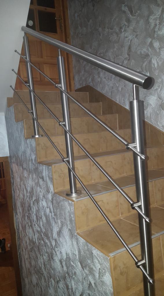 Polerowana, metalowa poręcz schodowa z trzema poziomami prętów, zamontowana na brązowych, ceramicznych schodach wewnątrz budynku, z widoczną dekoracyjną fakturą ściany w odcieniach szarości i drzwi...
