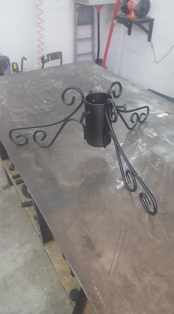 Czarny, metalowy stojak choinkowy z ozdobnymi, zakręconymi nogami, umieszczony na roboczym stole w warsztacie, widoczne ślady po obróbce metalu.