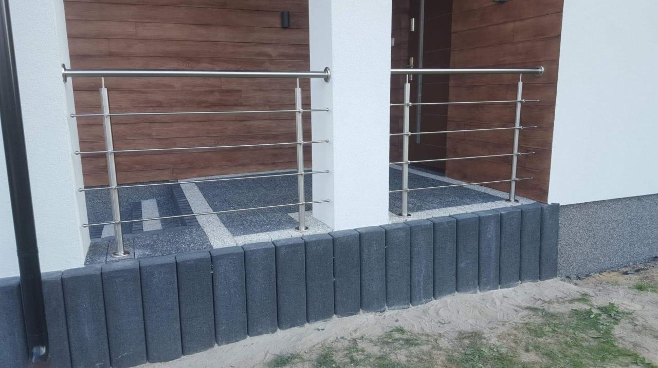 Nowoczesna balustrada ze stali nierdzewnej z poziomymi prętami, zamontowana na tarasie z szarej kostki brukowej, na tle elewacji z drewnianymi panelami.