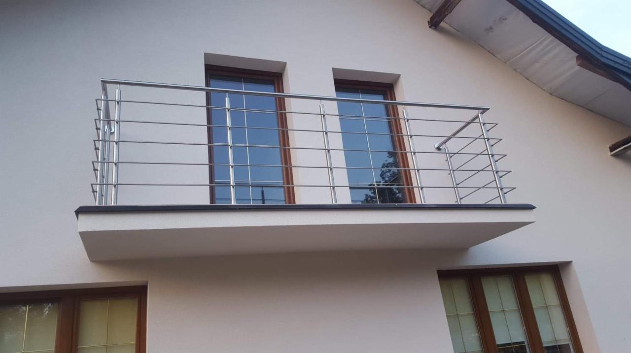 Balkon z balustradą ze stali nierdzewnej, widok z dołu, jasna elewacja budynku z oknami o brązowych ramach.