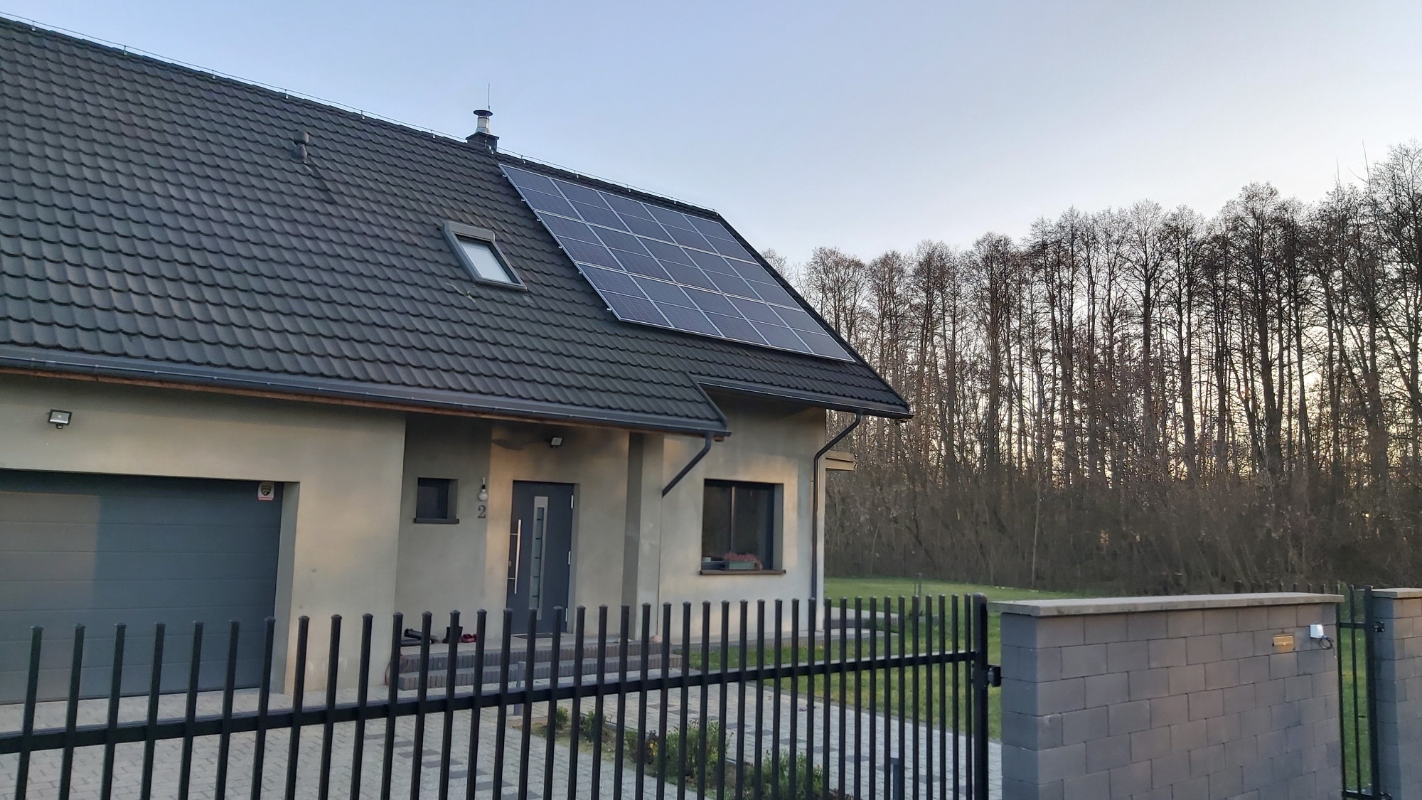 Dom jednorodzinny z szarej cegły z ciemną dachówką i panelami fotowoltaicznymi na dachu, ogrodzony czarnym metalowym płotem na tle drzew bez liści.