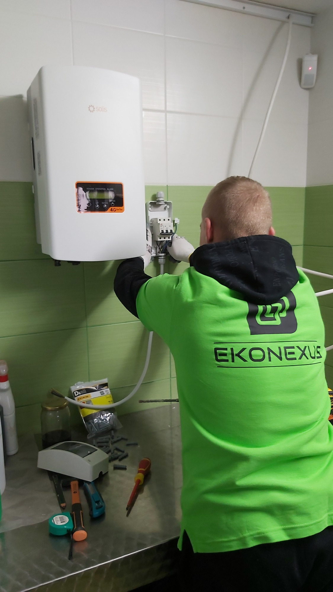 Instalator w zielonej koszulce z logo firmy EKONEXUS podłącza inwerter Solis 4G Series na ścianie z zielonymi płytkami, na stole leżą narzędzia i kołki montażowe.