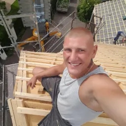 Uśmiechnięty mężczyzna na dachu w trakcie budowy więźby dachowej, widoczny dźwig i pracownik w oddali. Dach pokryty płytami OSB.