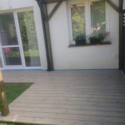 Stolarnia Skalin Tomasz Dolat - Jasnoszary taras z desek kompozytowych przylegający do białego domu z oknem, obok czarne drewniane ogrodzenie z prześwitami, widoczne kwiaty w doniczkach na parapecie