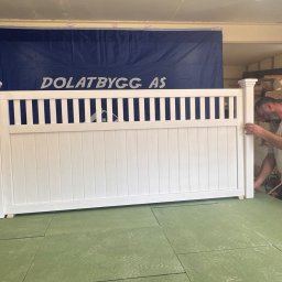 Stolarnia Skalin Tomasz Dolat - Białe, drewniane przęsło ogrodzeniowe w warsztacie, sprawdzane przez pracownika na tle reklamowego baneru firmy i półek z drewnem.