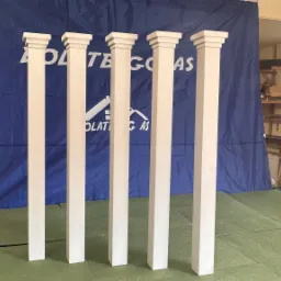 Pięć białych, kwadratowych kolumn z kapitelami ustawionych na zielonej powierzchni w warsztacie na tle niebieskiej plandeki.