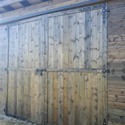 Rustykalne, dwuskrzydłowe drewniane wrota z ozdobnymi, kutymi zawiasami i zasuwką, wbudowane w ścianę budynku o drewnianej elewacji.