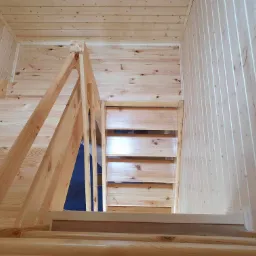 Wnętrze domu wykończone drewnem: schody z jasnego drewna prowadzące na górę, drewniane ściany i sufit. Widoczny fragment niebieskiej folii budowlanej.