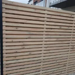 Nowoczesne ogrodzenie z poziomych desek z jasnego drewna, z czarnym słupkiem po lewej stronie, na tle budynku z balkonem.