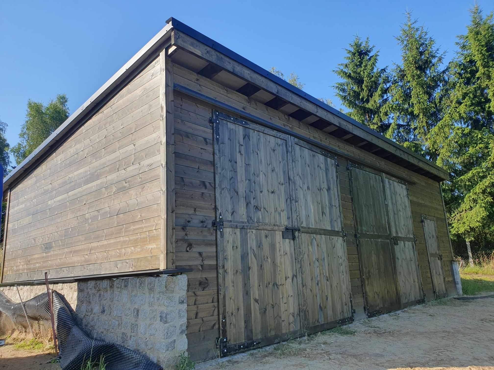 Drewniana konstrukcja z przesuwnymi drzwiami, osadzona na kamiennej podmurówce, widoczna z niskiej perspektywy w słoneczny dzień. Elementy metalowe mocujące drzwi. W tle drzewa iglaste.