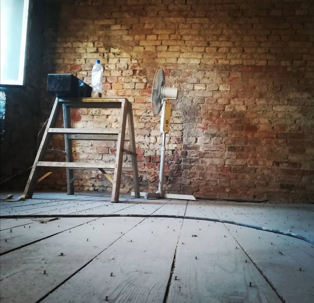 Pokój w trakcie remontu: stara ceglana ściana, drewniana podłoga z wystającymi gwoździami, drabina z butelką wody i głośnikiem, obok wentylator.