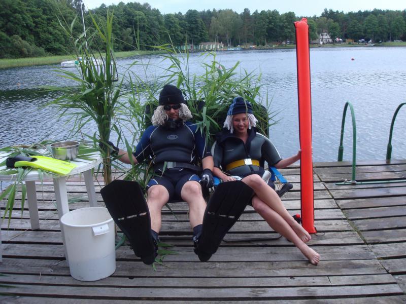 Dwoje nurków w strojach, siedzących na pomostach nad jeziorem, obok trzcin i sprzętu nurkowego, z dmuchanym czerwonym bojami.