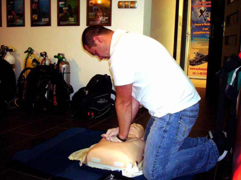 Mężczyzna w białej koszulce i jeansach ćwiczy resuscytację krążeniowo-oddechową na manekinie na tle sprzętu do nurkowania i plakatów.